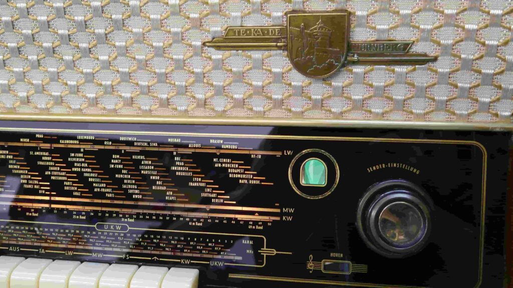 Nahaufnahme eines TeKaDe Röhrenradios mit sichtbarem Lautsprechergitter, Stoff weiß/gelb, Markenemblem und Skala mit Frequenzanzeige, Magisches Fächerauge sowie Sender Einstelldrehknopf.